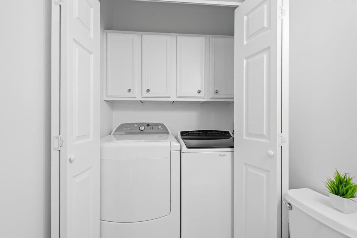 A laundry room with a front-loading washer and dryer, white cabinets above, and a small plant on the right.