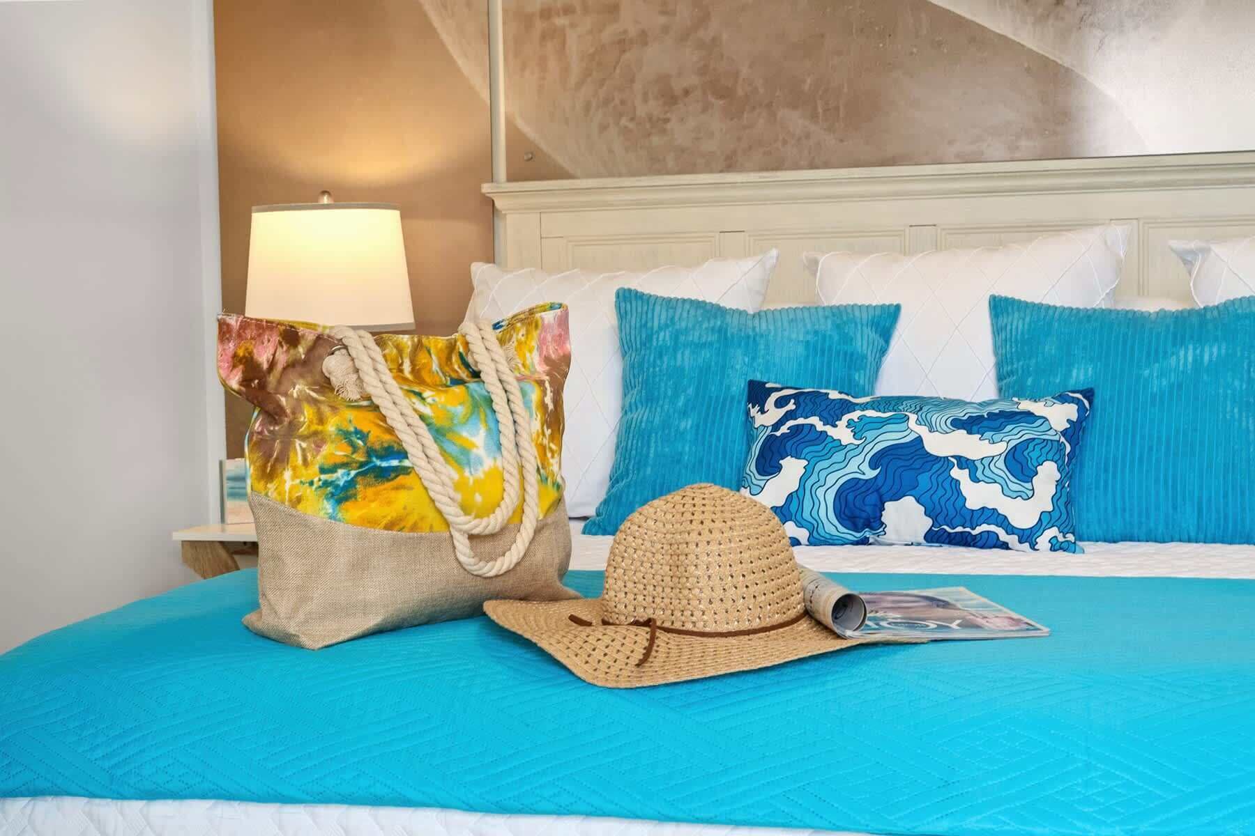 A bed with a blue quilt, a straw hat, a woven bag, and two patterned pillows.