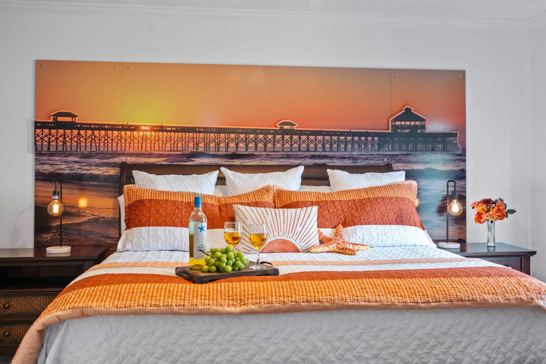 En säng med en tavla som föreställer en solnedgång, orange sängkläder och ett brickbord med druvor och en flaska.