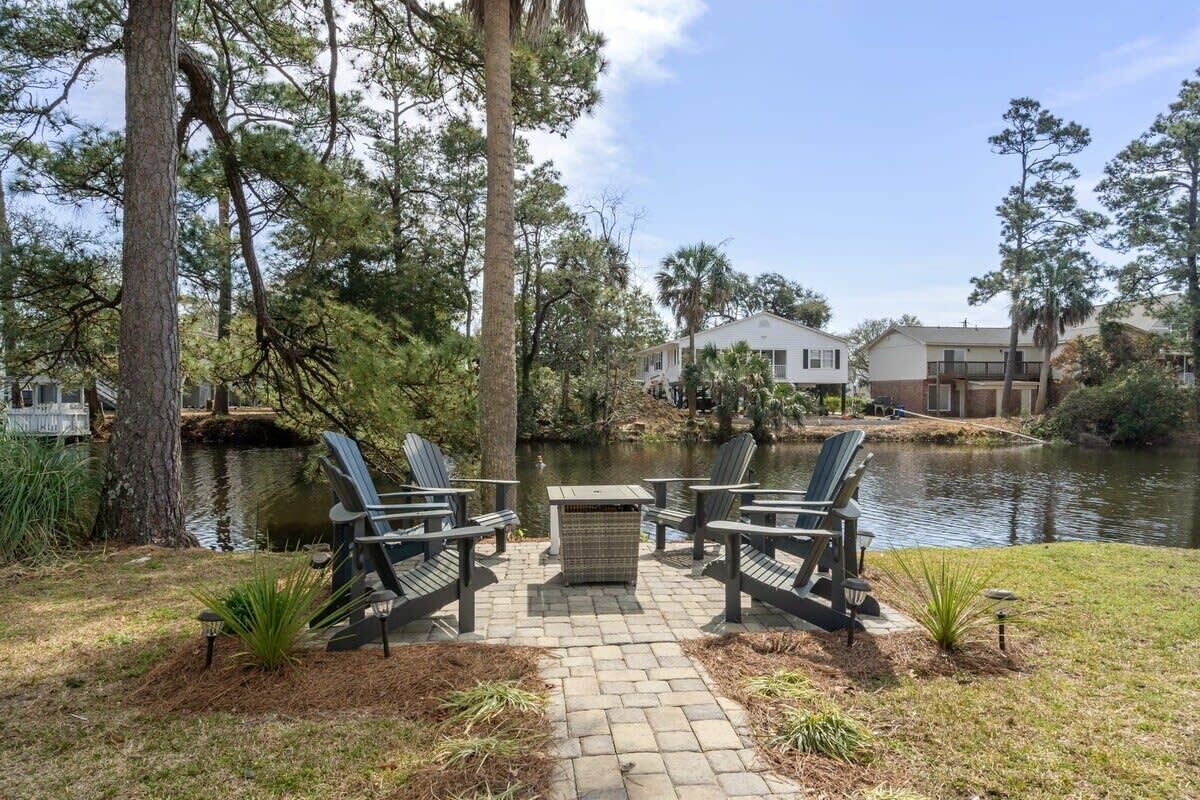 A patio with Adirondack chairs and a table by a pond, surrounded by trees and a house in the background.