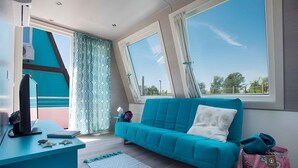 A modern room with a teal sofa, a desk with a computer, and large windows offering a view of the outdoors. - Holiday home with air conditioning and terrace (Lignano Sabbiadoro)