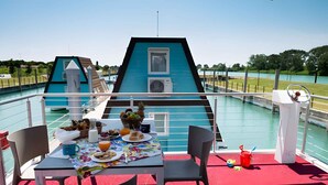 A dining area with a table set for two, overlooking a body of water, a boat, and a grassy area. - Holiday home with air conditioning and terrace (Lignano Sabbiadoro)