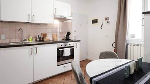 Cocina pequeña con armarios blancos, estufa y pileta. Se observa una mesa de comedor con una silla y un radiador al fondo.