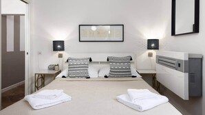Habitación de hotel con cama, dos almohadas, mesita de noche, lámpara, un cuadro en la pared y un radiador.