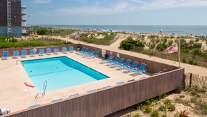 Zona de piscina con sillas azules y una bandera, con vista a una playa con gente y sombrillas.