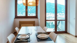 Een eetruimte met een tafel voor vier personen, grote ramen met uitzicht op een heuvelstad, en een hanglamp boven de tafel.
