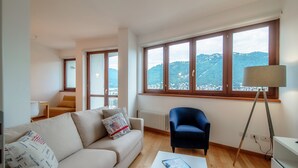 Een moderne woonkamer met een bank, een stoel, een bijzettafeltje en een groot raam met uitzicht op een bergachtig landschap.
