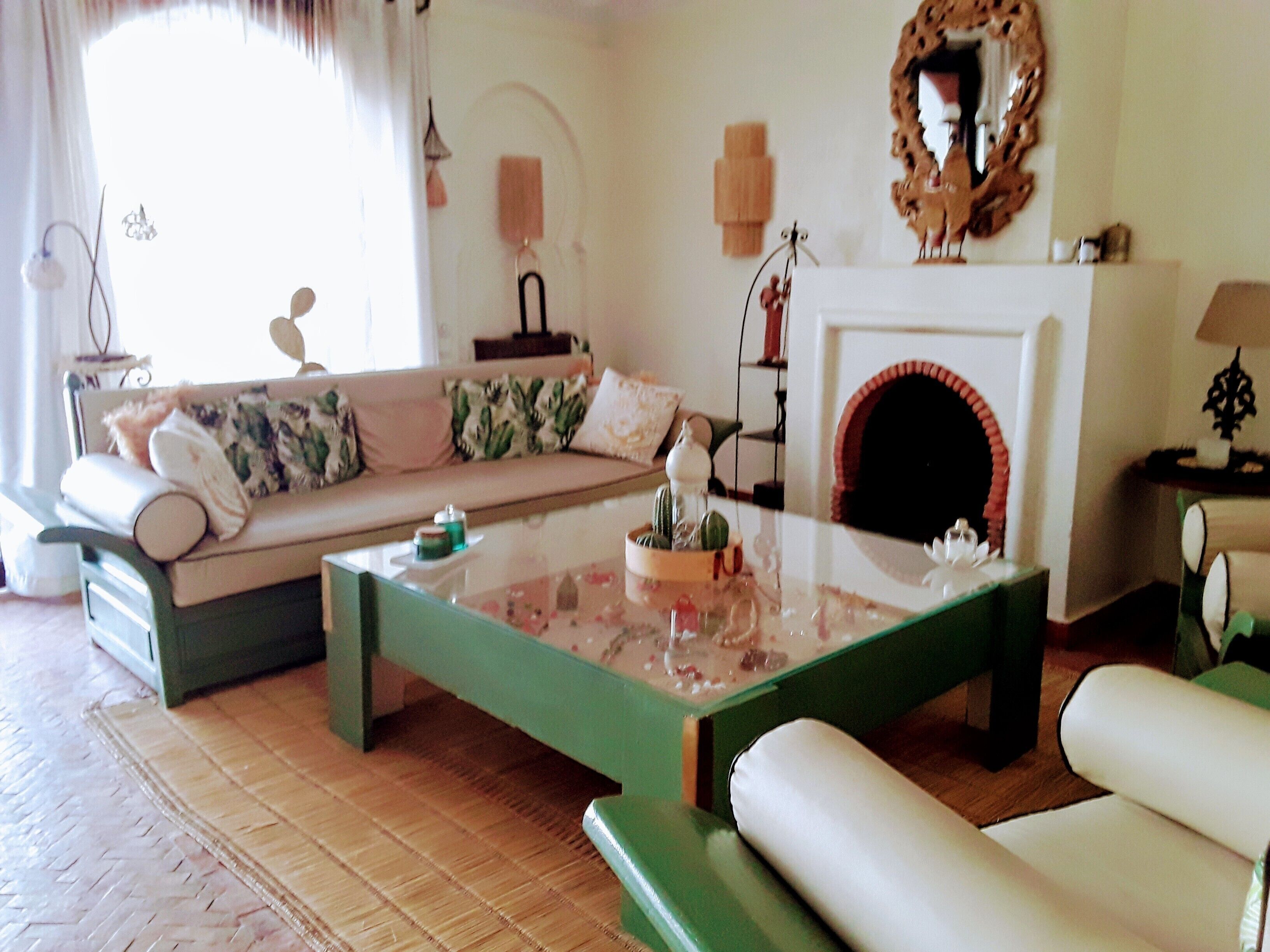 A cozy living room with a green coffee table, a white sofa with floral cushions, a fireplace, and a mirror on the wall.
