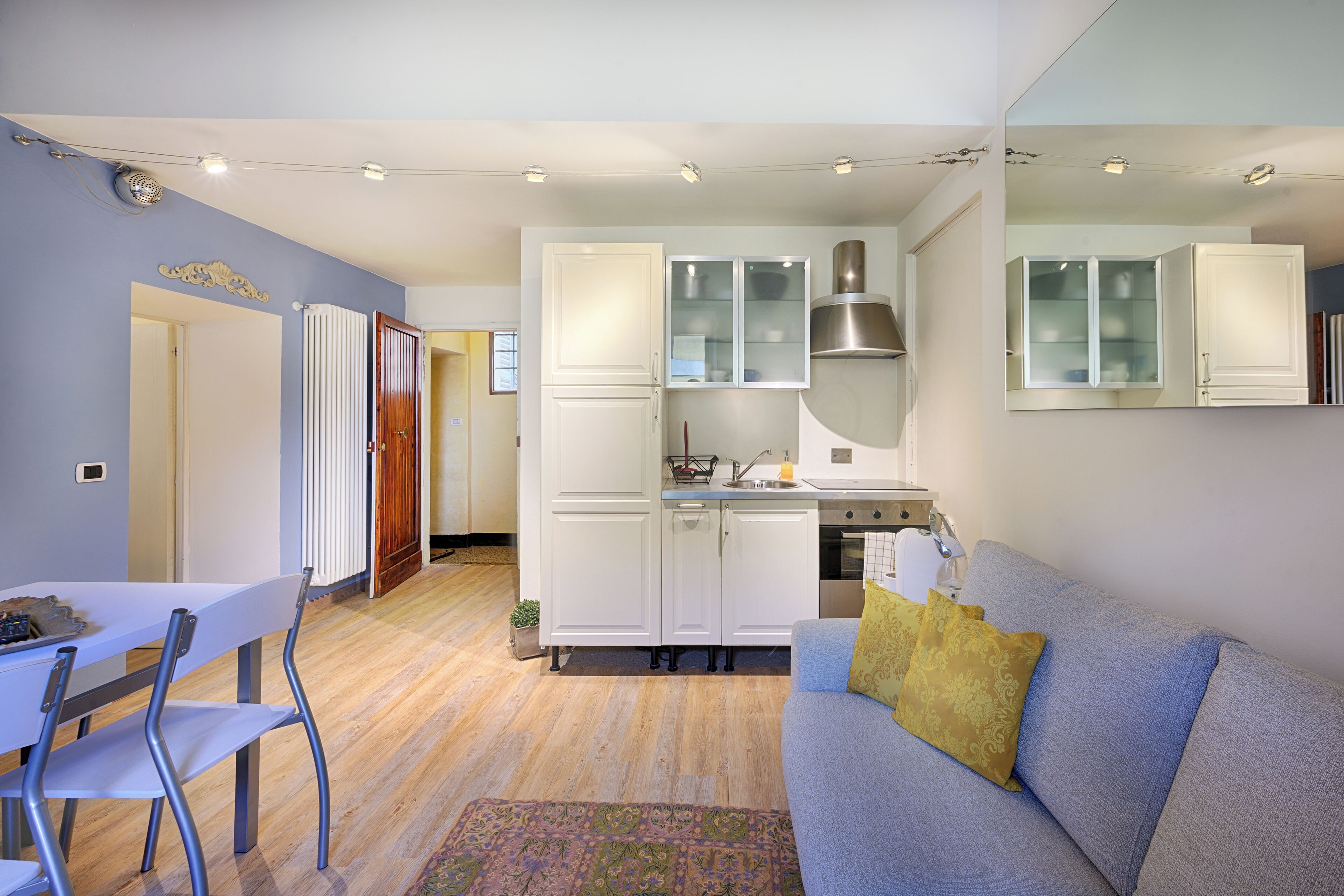 A modern kitchen with white cabinets, a built-in stove, and a microwave. There is a dining area with a table and chairs, and a living room with a sofa and a patterned rug.