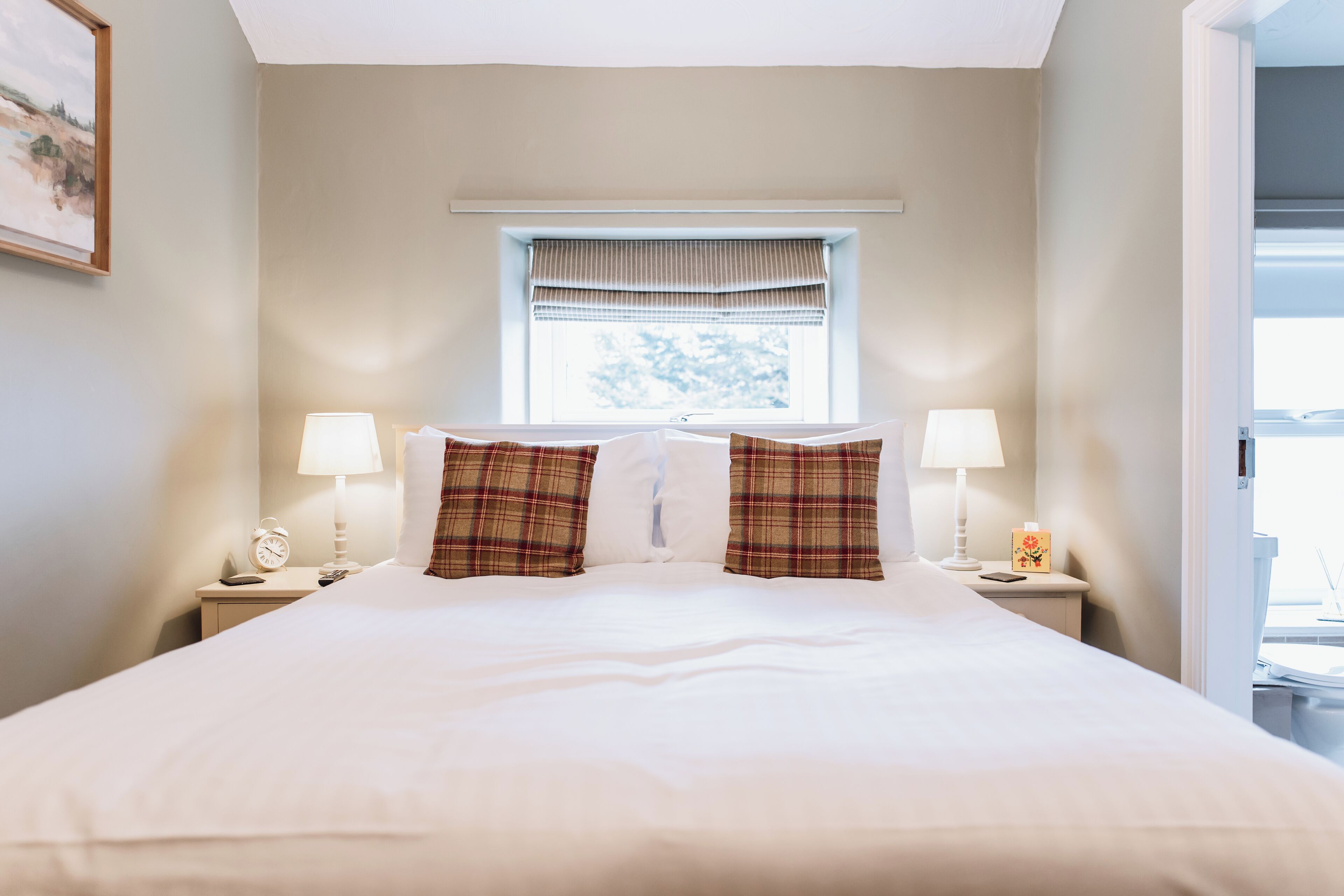 A neatly made bed with plaid pillows, two bedside lamps, a clock, and a window with blinds.