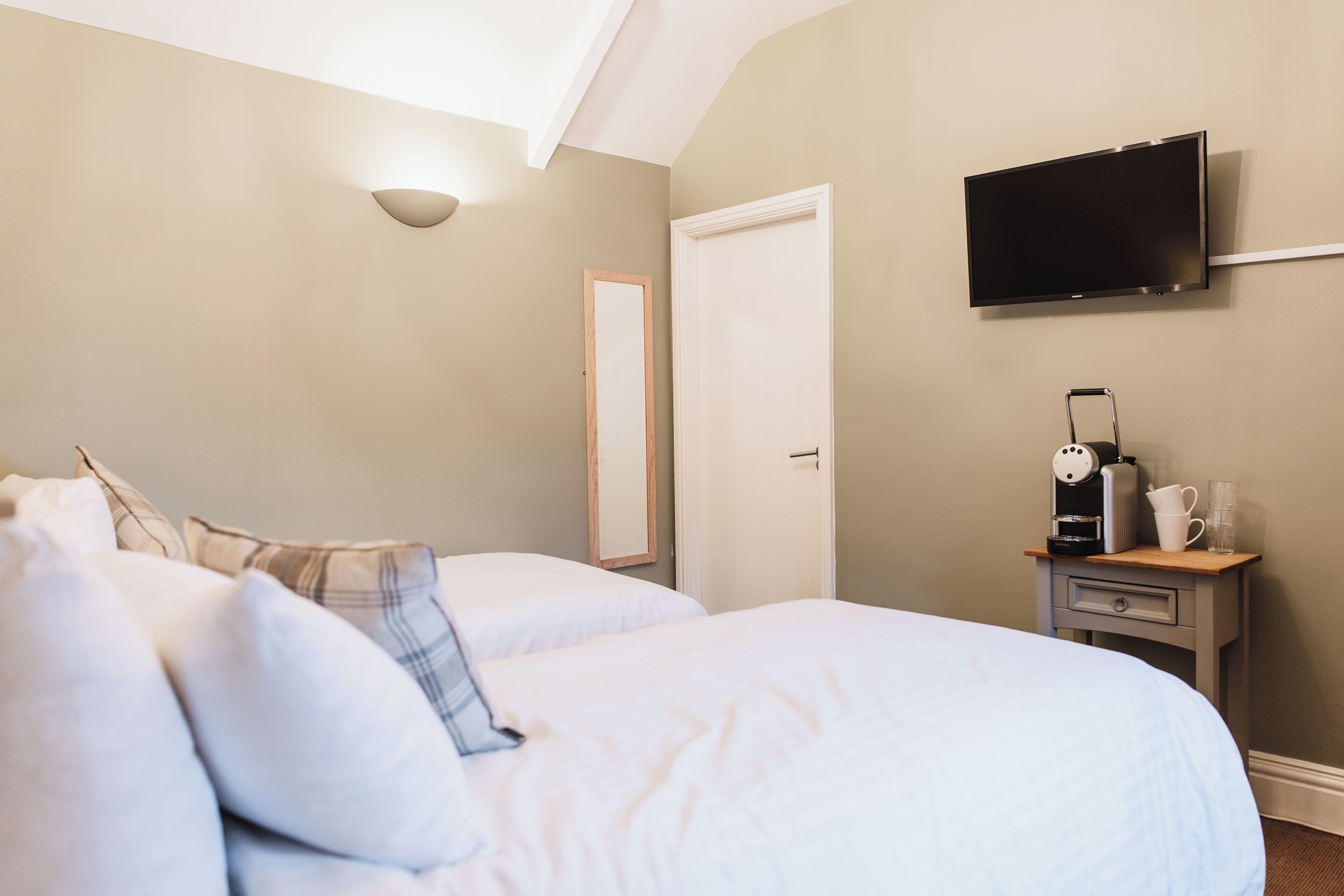 Una habitación de hotel con dos camas, un televisor de pantalla plana montado en la pared, una mesita de noche con cafetera y un espejo.
