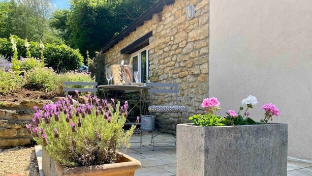 A stone building with a patio area featuring a table and chairs, surrounded by potted plants and flowers. - Little Innsacre (Shipton Gorge)