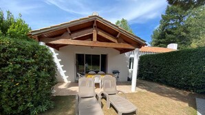 A patio with a table and chairs, surrounded by a hedge and a building with a sloped roof. - Cozy 2-BR House with Terrace, Near Forest and Beaches, Sleeps 6 (Saint-Jean-de-Monts)