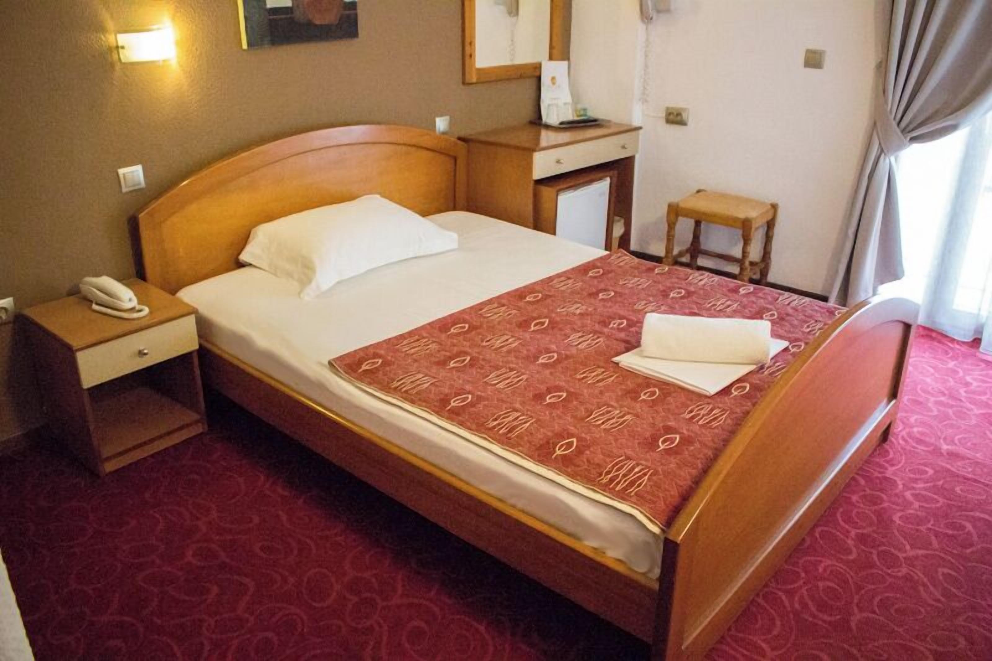 Habitación de hotel con cama de madera, mesita de noche con teléfono, escritorio pequeño con lámpara y ventana con cortinas.