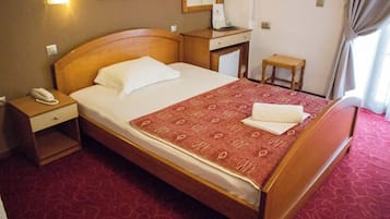 Habitación de hotel con cama de madera, mesita de noche con teléfono, escritorio pequeño con lámpara y ventana con cortinas.