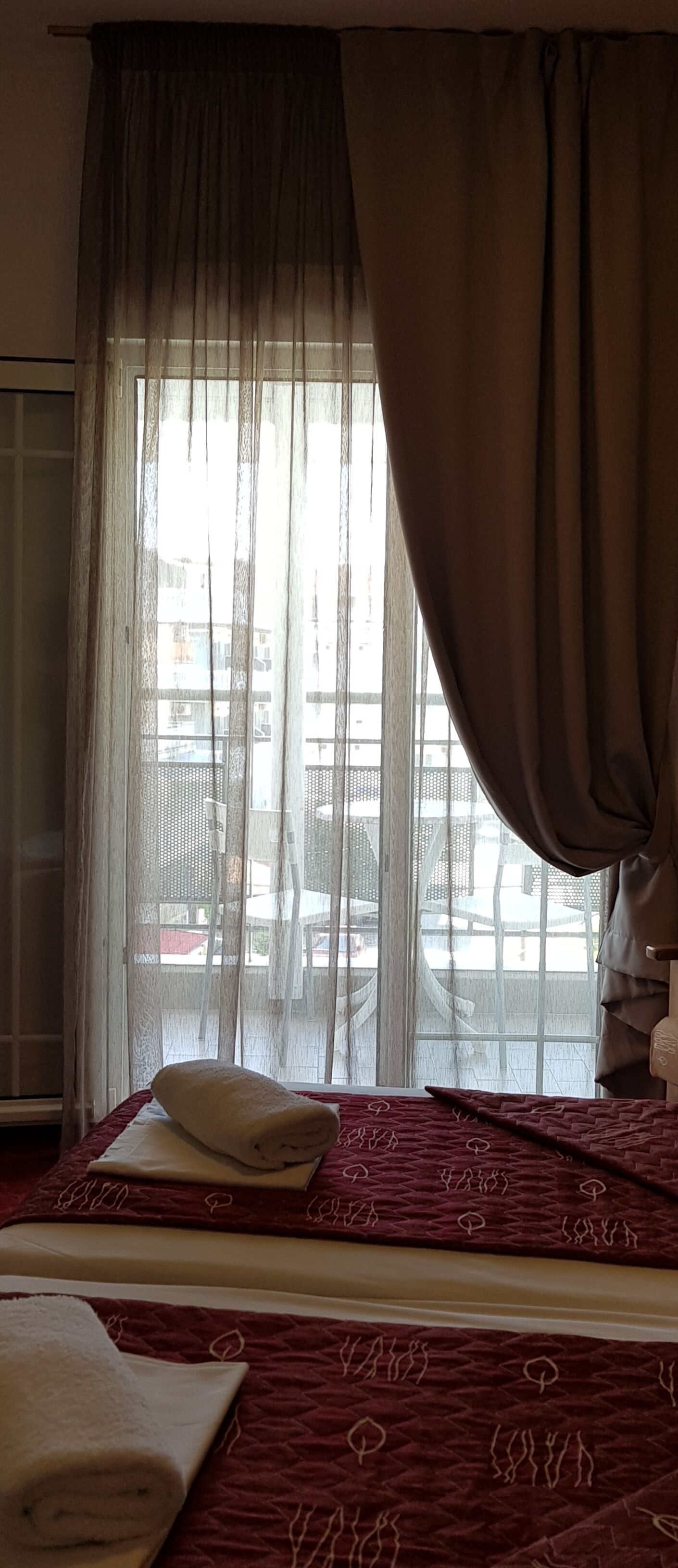 Quarto de hotel com cama, janela com cortinas leves e vista para prédios.