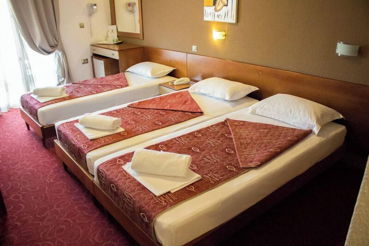 A hotel room with three single beds, each with a red patterned bedspread and white pillows. There is a wooden headboard, a bedside table with a lamp, and a small desk with a telephone.