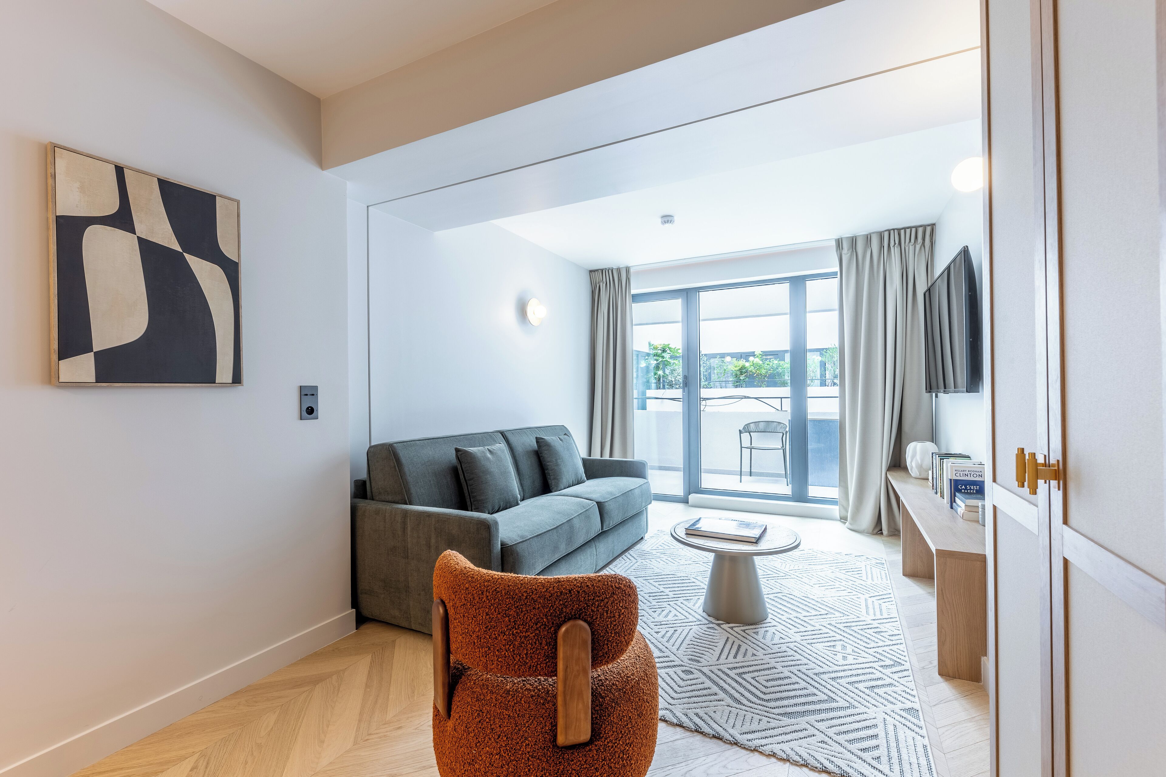 Ein modernes Wohnzimmer mit einem grauen Sofa, einem braunen Sessel und einem kleinen Couchtisch. Ein großes abstraktes Gemälde hängt an der Wand, und ein Fernseher ist dort montiert.