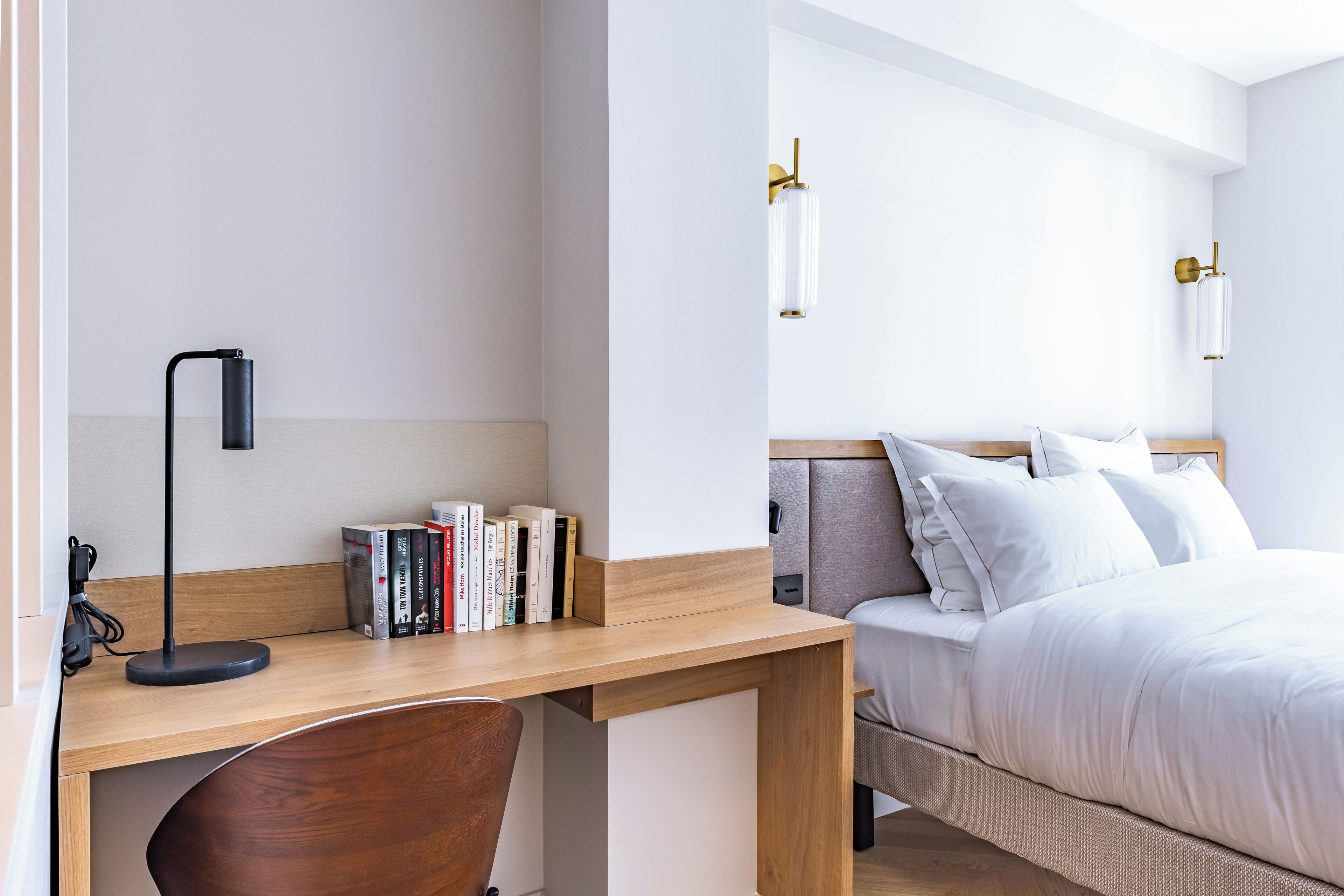 Een moderne hotelkamer met een houten bureau, een stoel, een bed met witte lakens en twee wandlampen.