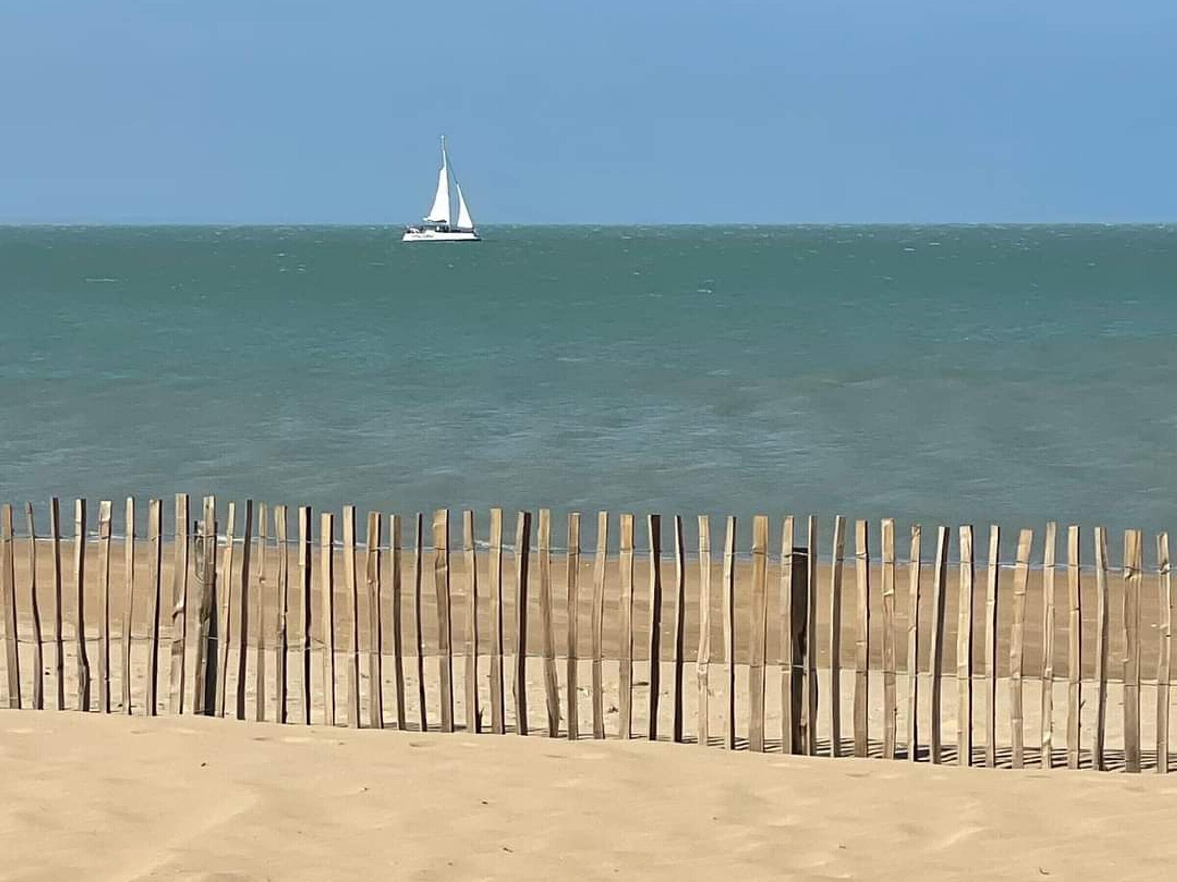 A sandy beach with a wooden fence in the foreground and a sailboat on the water in the background.