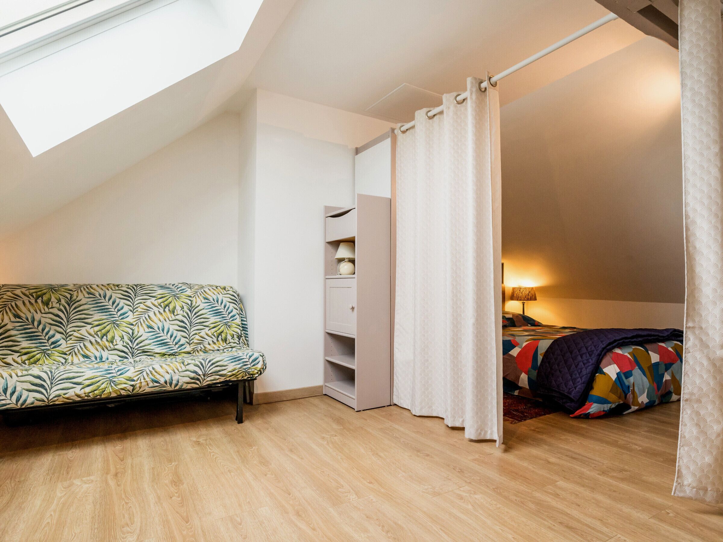 A cozy attic bedroom with a sloped ceiling, a sofa, a bed with a colorful quilt, and a window with a curtain.