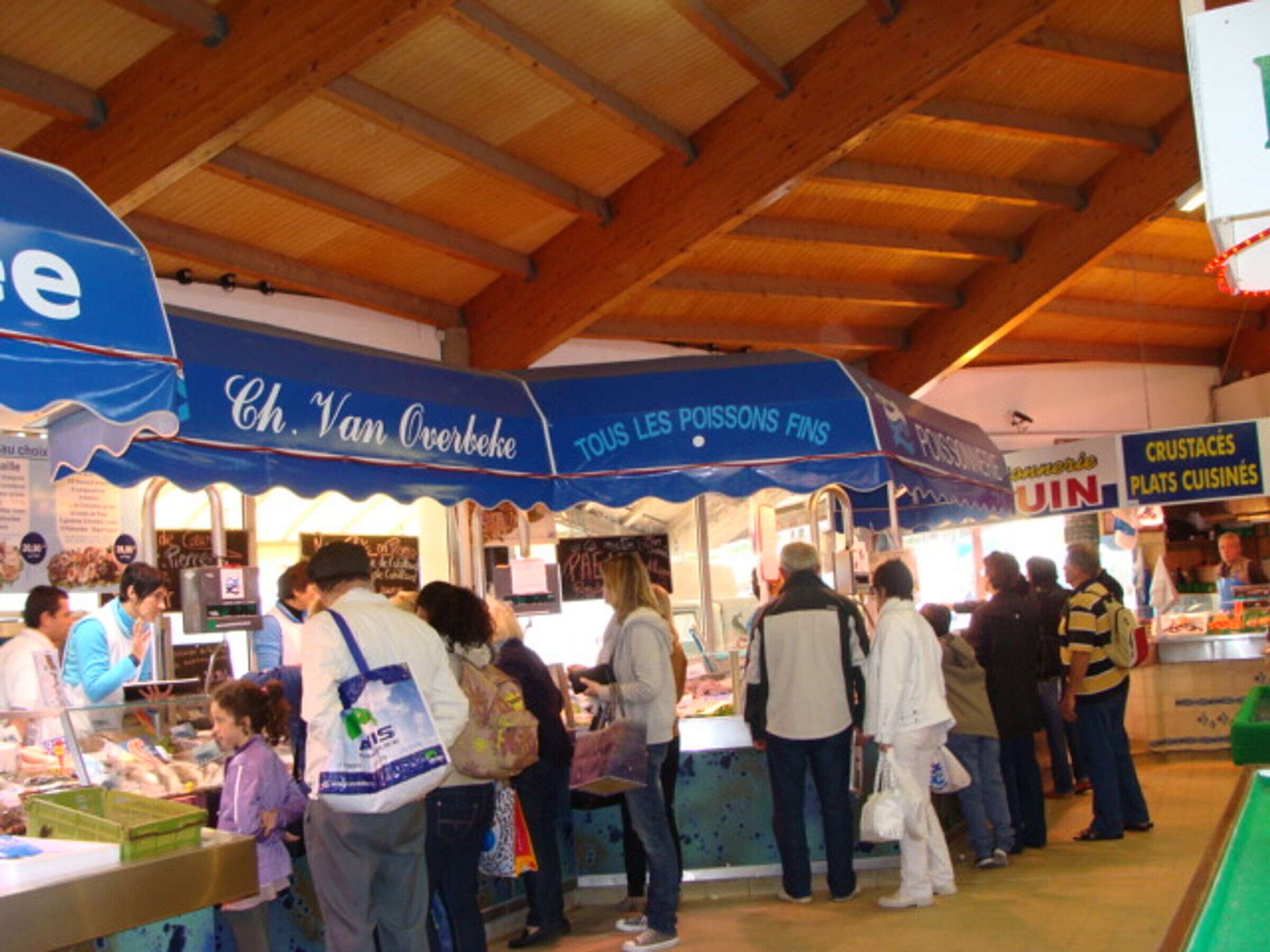 A market stall with a blue awning labeled "Ch. Van Overbeke" and another stall with a sign "TOUS LES POISSONS FINS".