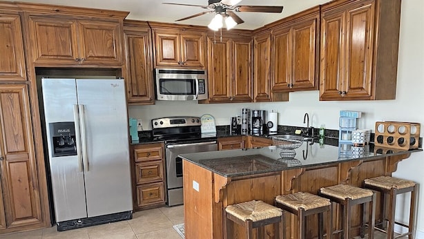 A kitchen with wooden cabinets, a stainless steel refrigerator, a microwave, a stove, and a bar area with stools.