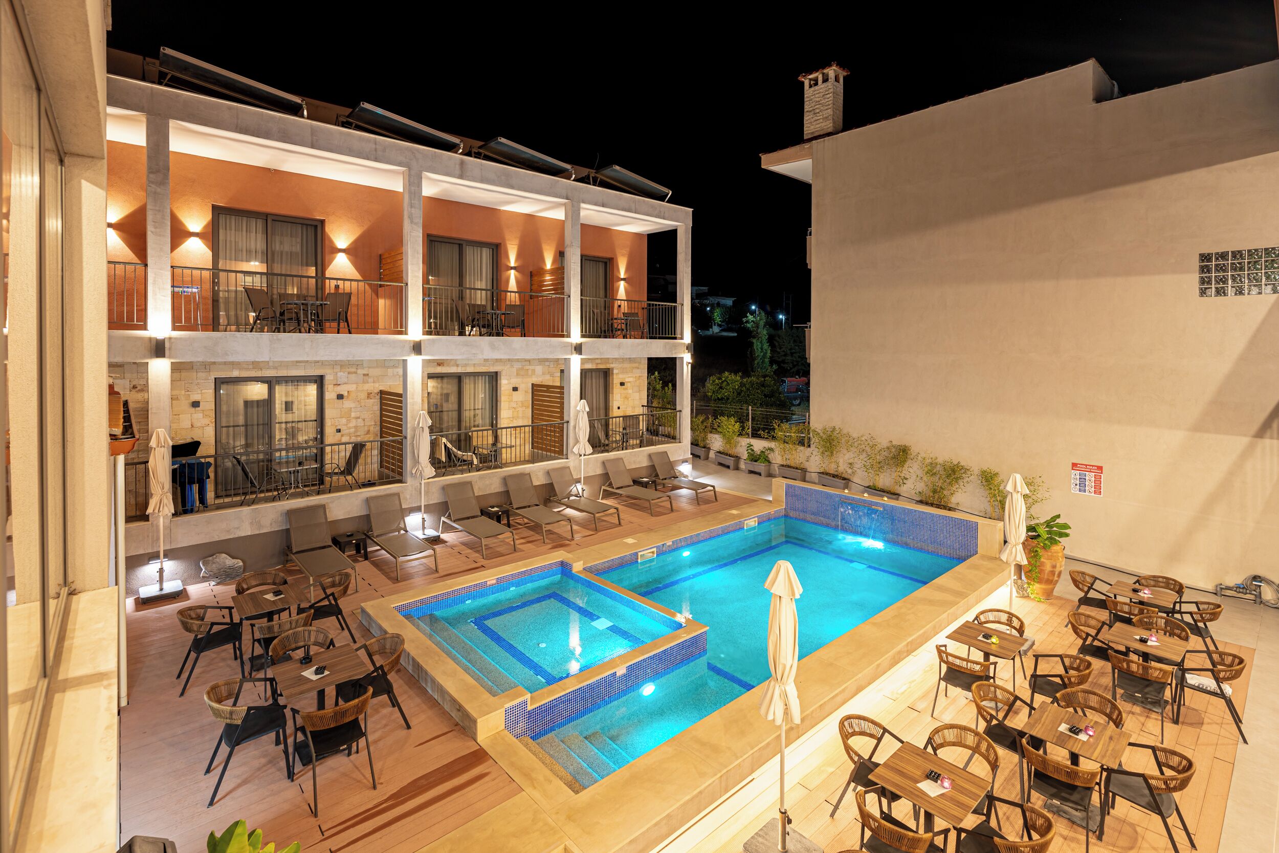 A hotel with a rooftop pool, outdoor seating, and a second-floor balcony with lounge chairs.