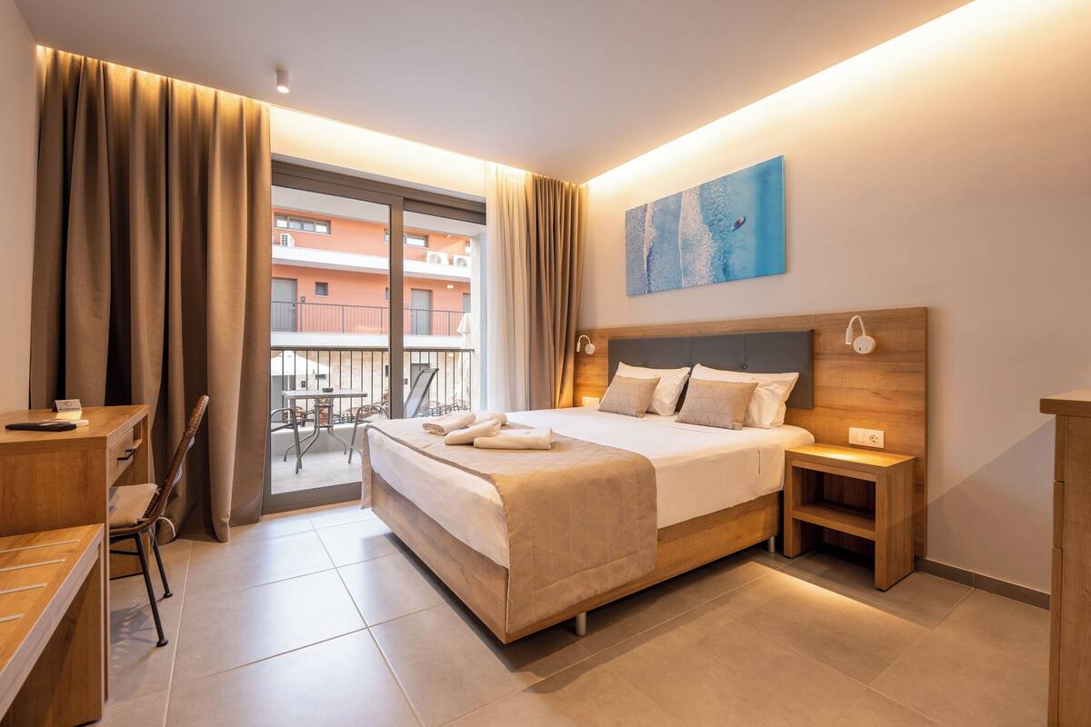 Habitación de hotel con cama, mesita de noche, silla, escritorio y vistas al balcón exterior.