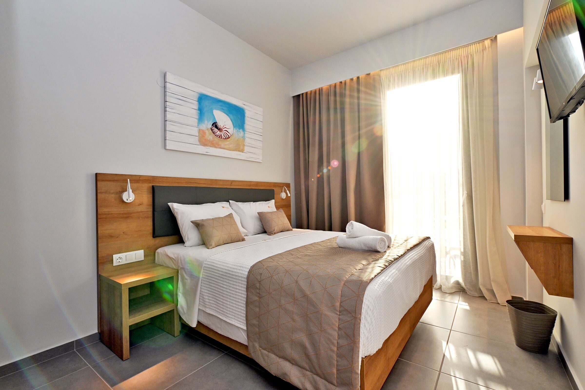 Habitación de hotel con cama, mesita de noche, televisor, una obra de arte, cortinas y estante en la pared.