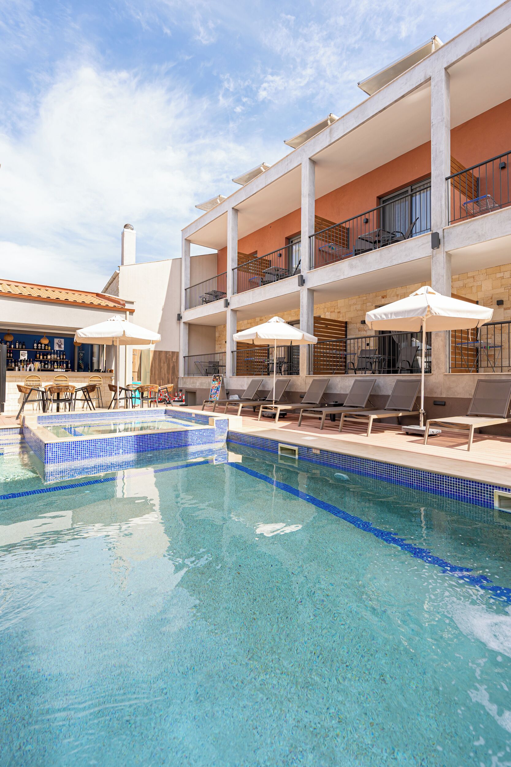 Zona de piscina amb gandules i ombrel·les, un bar i un edifici amb balcons.