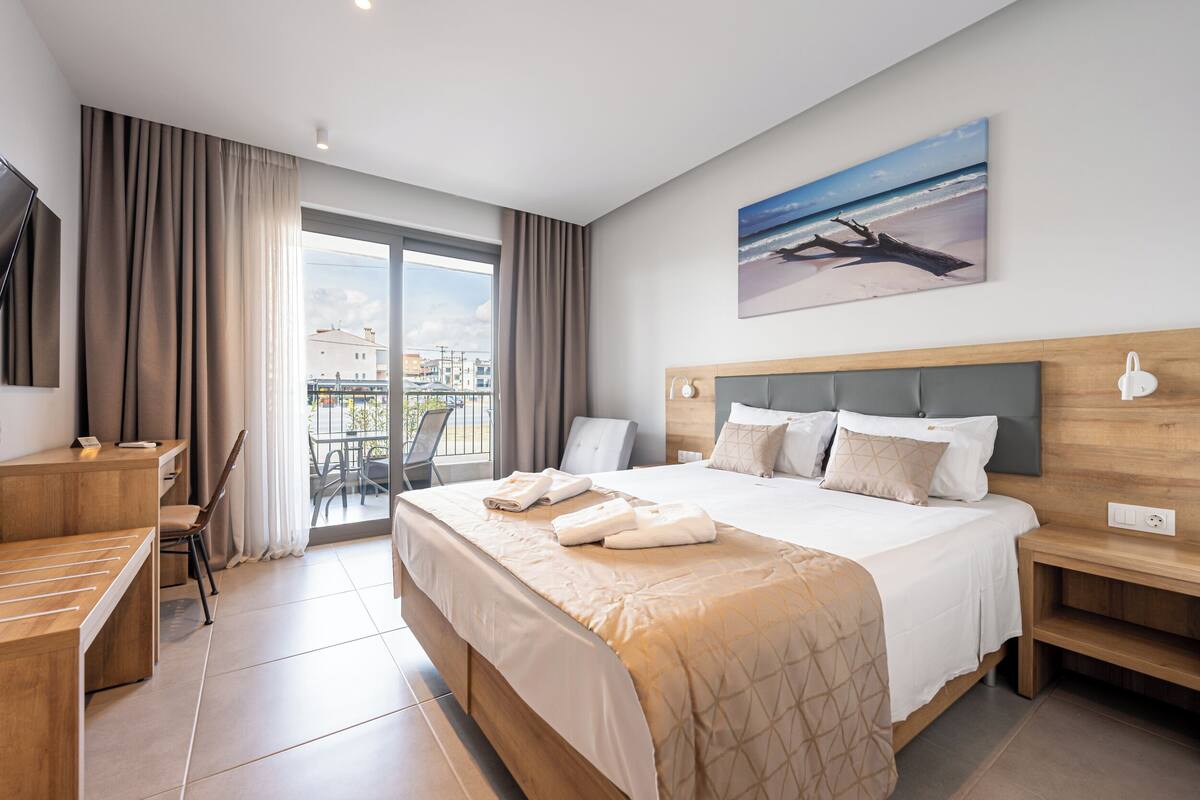 Habitación de hotel con cama, escritorio, silla, balcón con vistas y un cuadro en la pared.