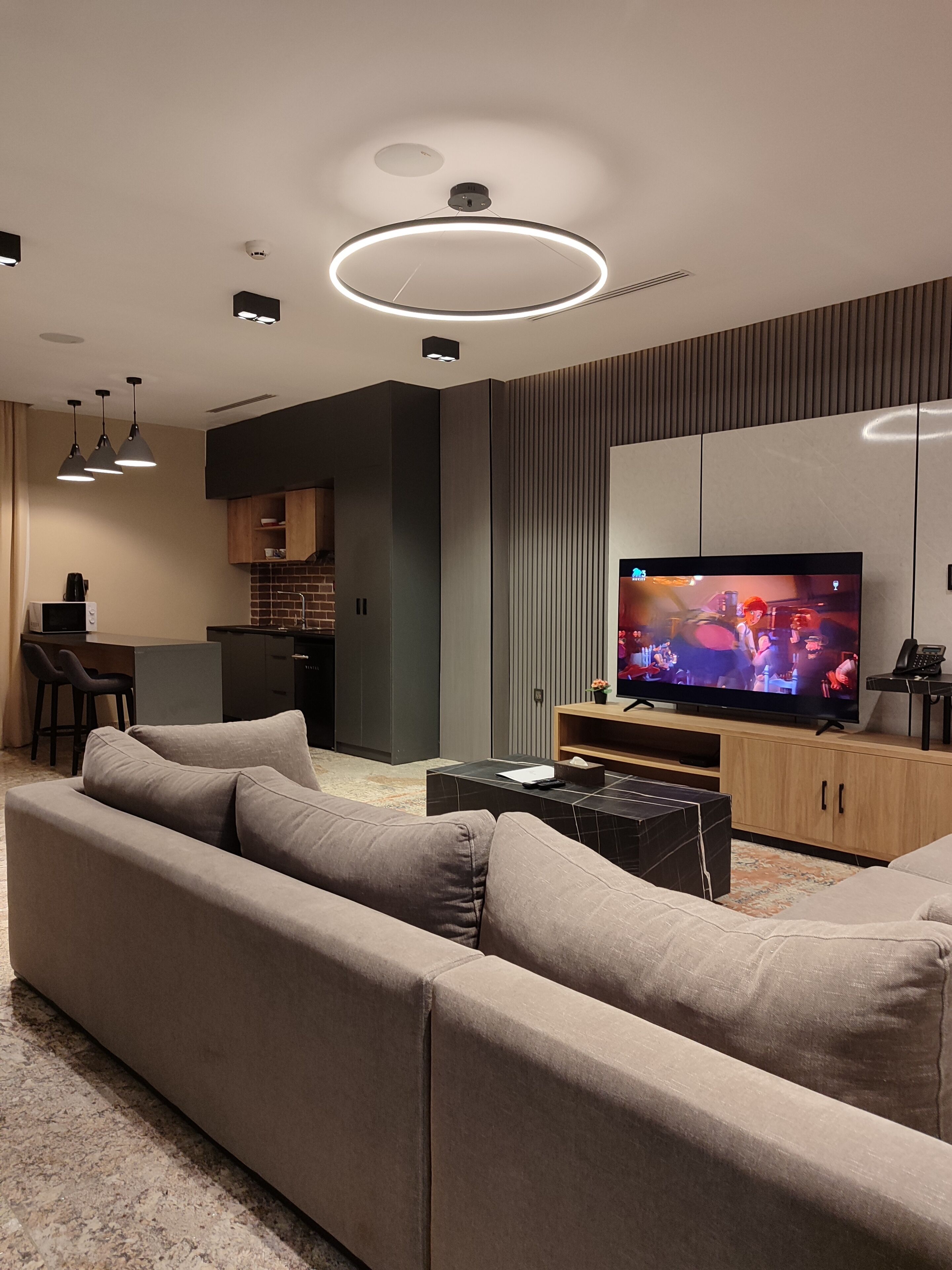 A modern living room with a large flat-screen TV, a sofa, a coffee table, pendant lights, and a kitchen area in the background.