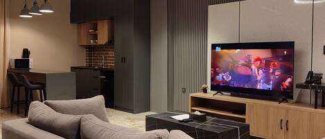 A modern living room with a large flat-screen TV, a sofa, a coffee table, pendant lights, and a kitchen area in the background.