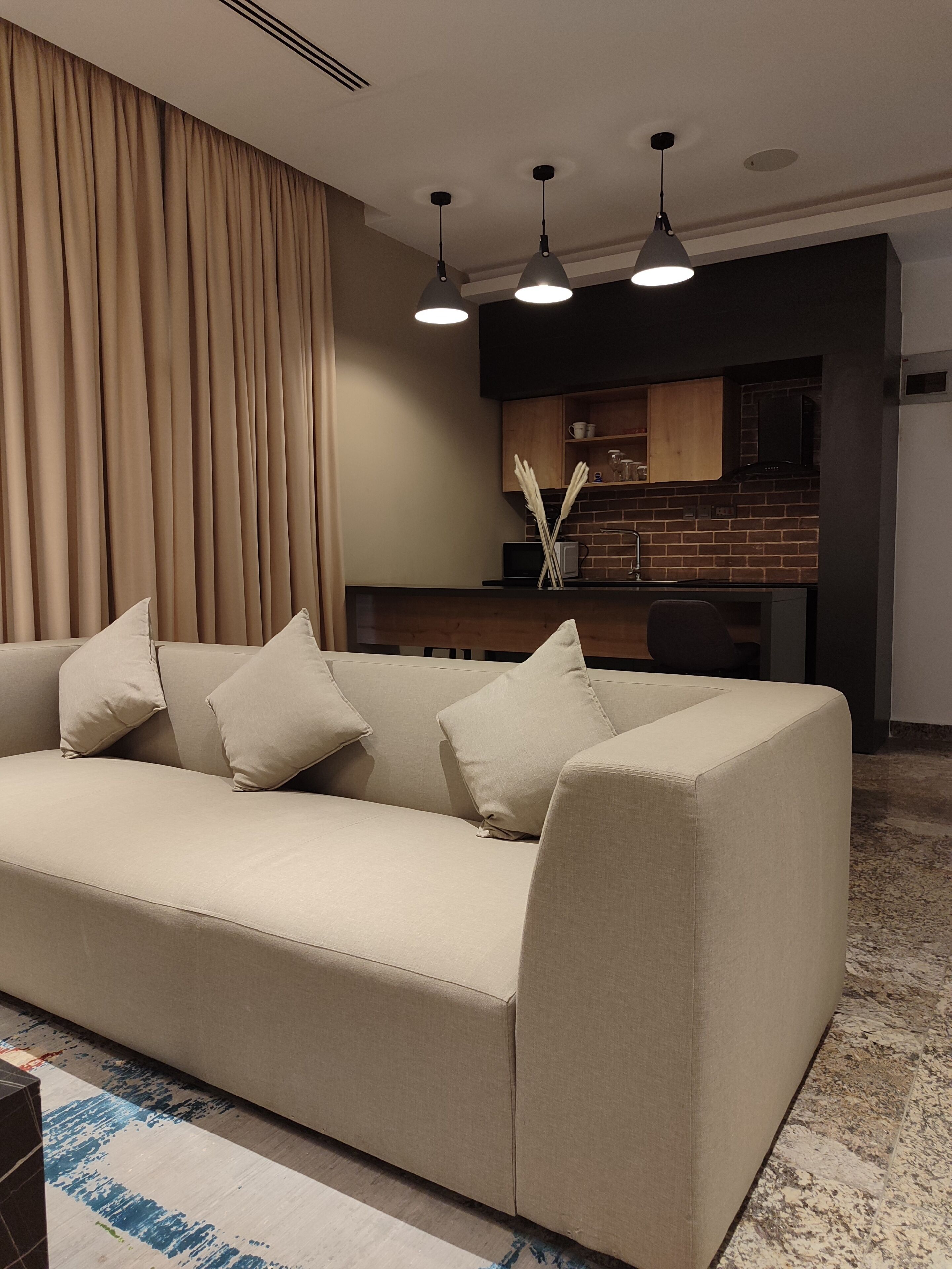 A modern living room with a beige sofa, a kitchen in the background, and a large window with curtains.