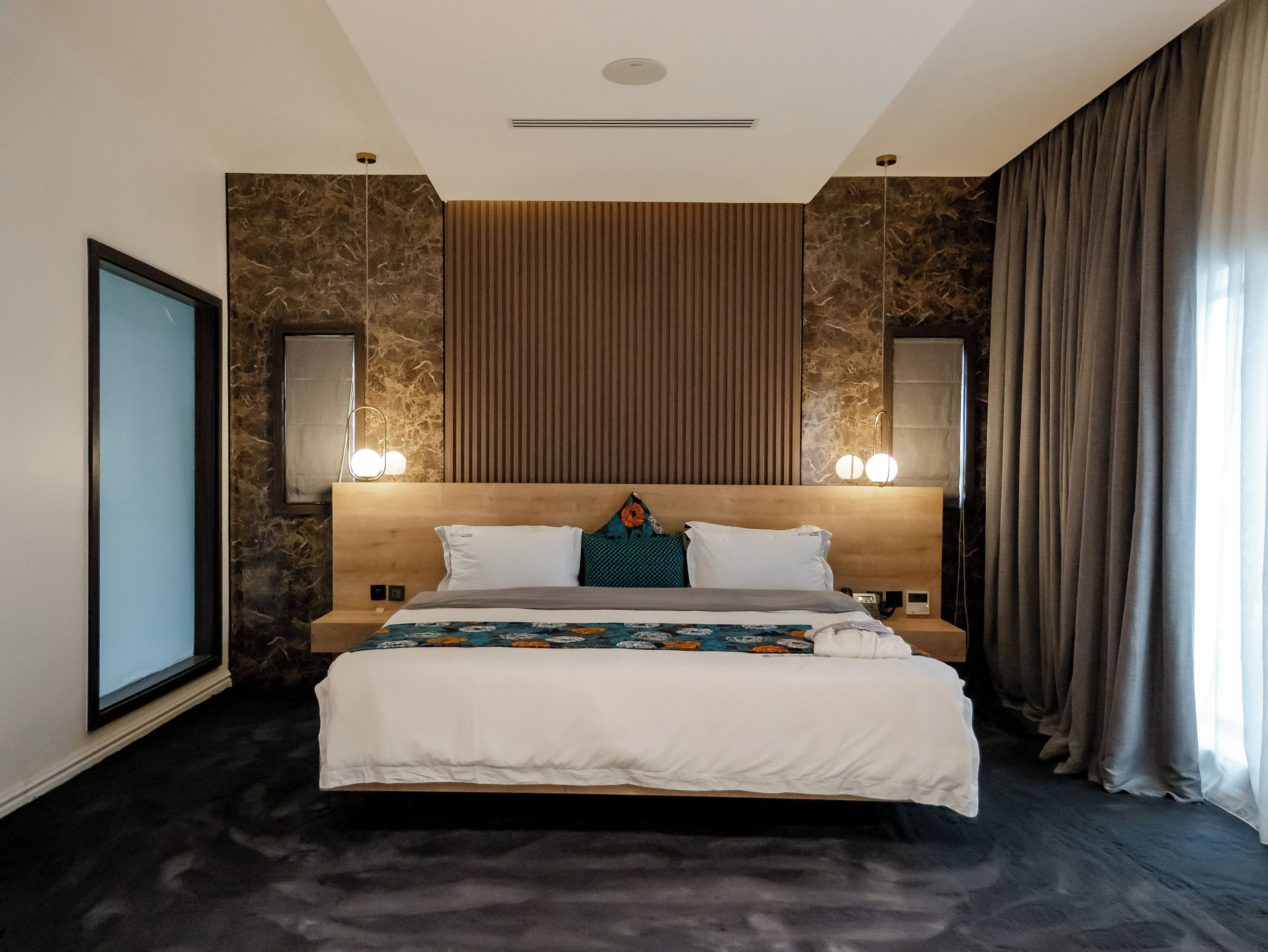 A modern hotel room with a large bed, a wooden headboard, a marble accent wall, and a large window with curtains.