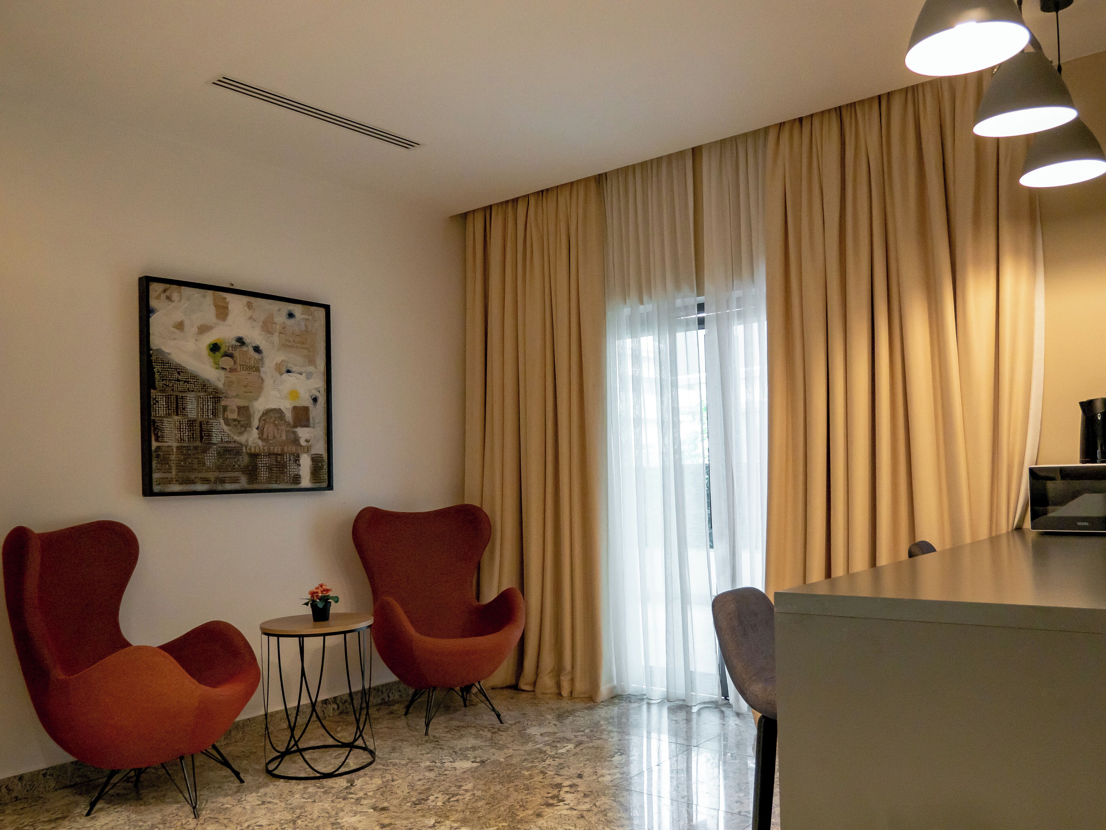A hotel room with a modern design, featuring two red chairs, a small table with a flower, a framed picture on the wall, and a large window with curtains.