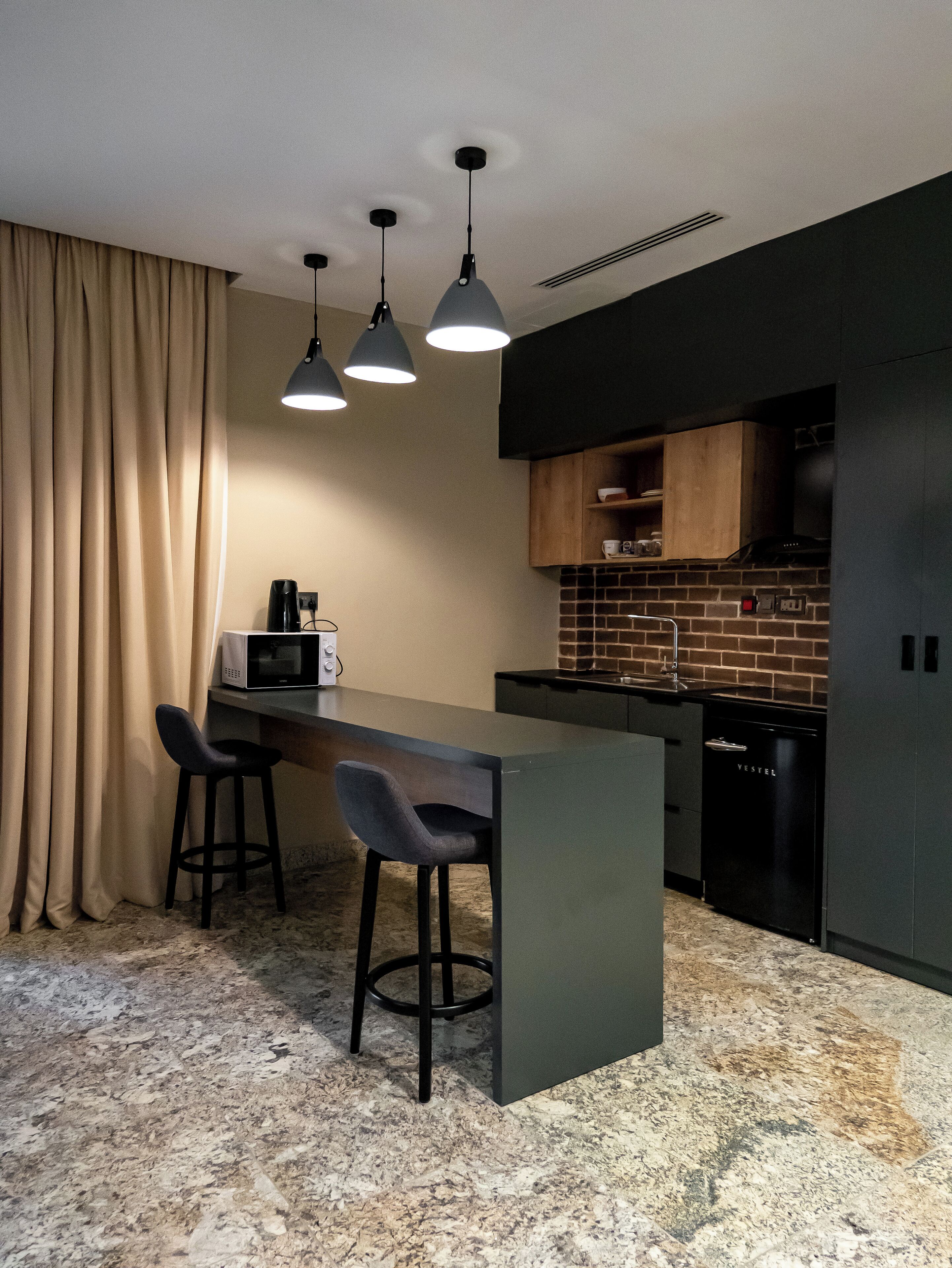 Cocina compacta con encimera oscura, microondas y lavamanos. Hay dos taburetes de bar y una lámpara colgante.