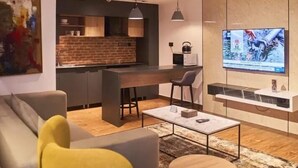 A modern living room with a sofa, coffee table, and TV. There is a kitchen area with a brick backsplash and a dining table. - BRICKSPOINT BOUTIQUE APARTHOTEL ASOKORO (Abuja)
