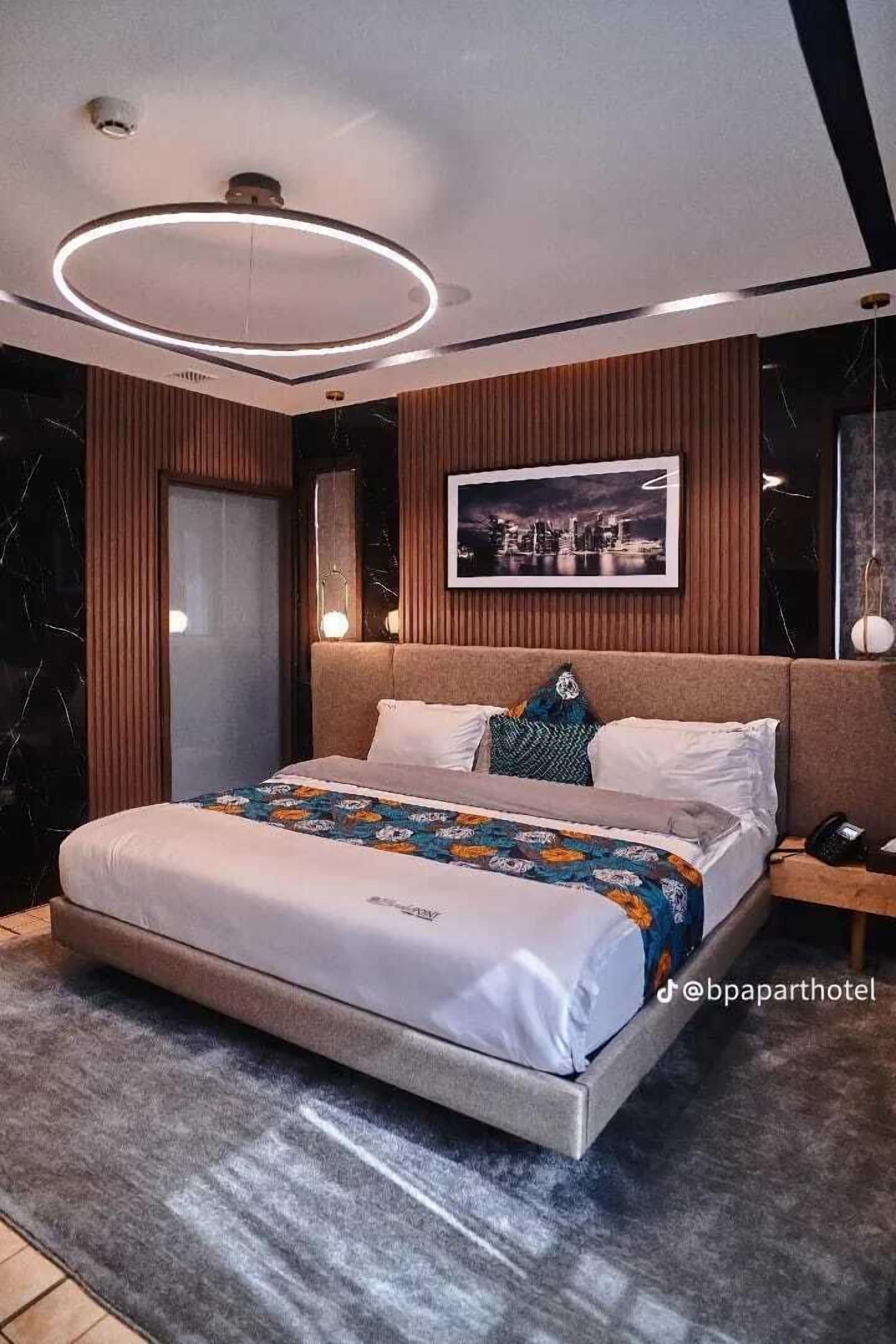 Une chambre d’hôtel moderne avec un grand lit, un luminaire rond au plafond, un tableau représentant un paysage urbain et un mur recouvert de panneaux de bois.