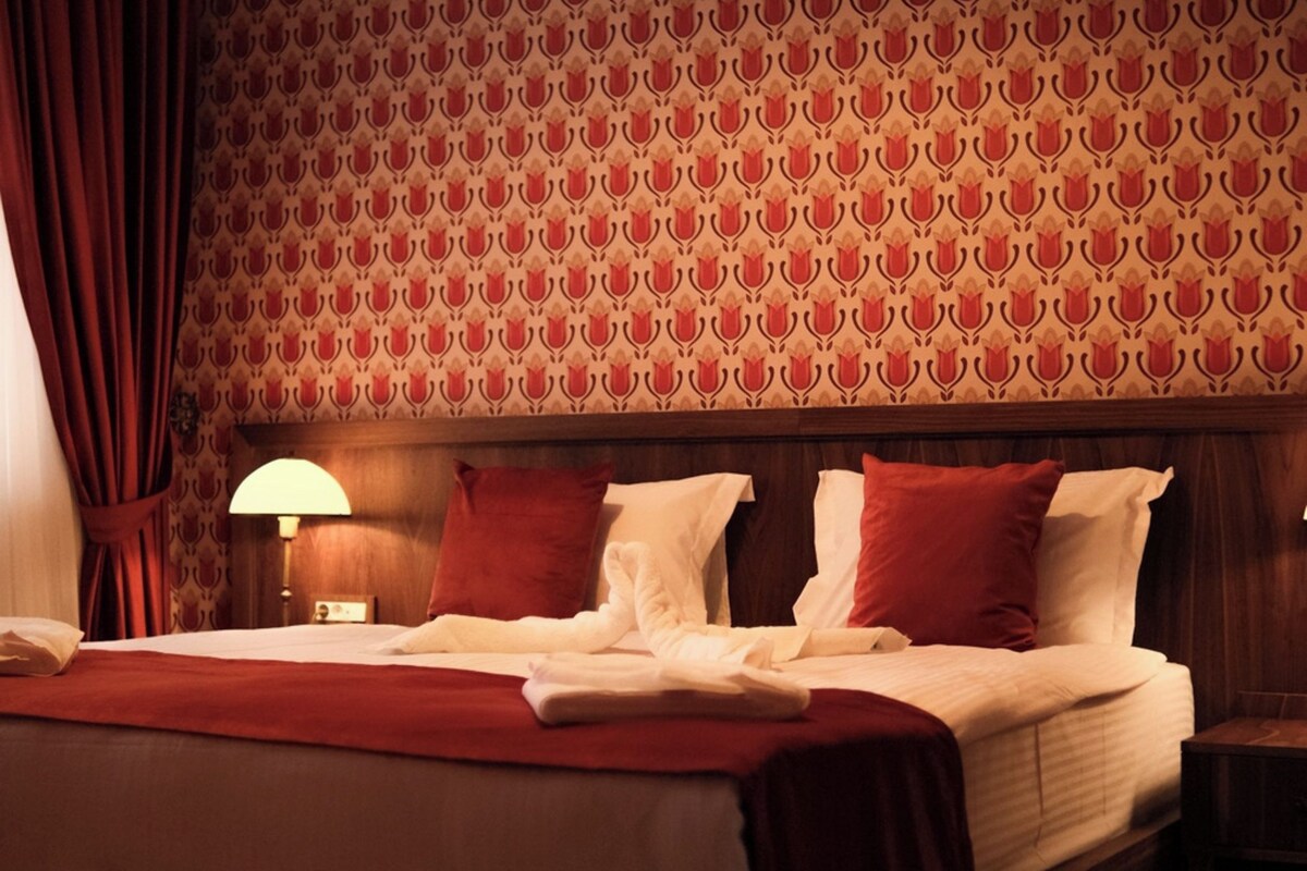 A hotel room with a large bed, red curtains, patterned wallpaper, and a nightstand with a lamp.
