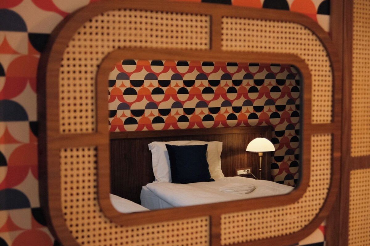 A room with a geometric patterned wallpaper, a bed with white linens, a dark blue pillow, and a bedside lamp.