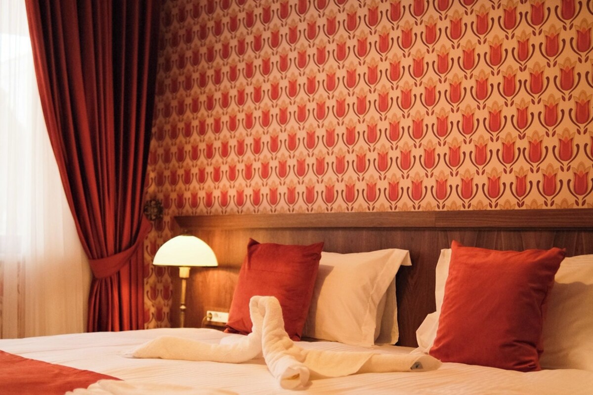 Hotelkamer met behang met patroon, een bed met rood-witte beddengoed, een lamp en een raam met rode gordijnen.