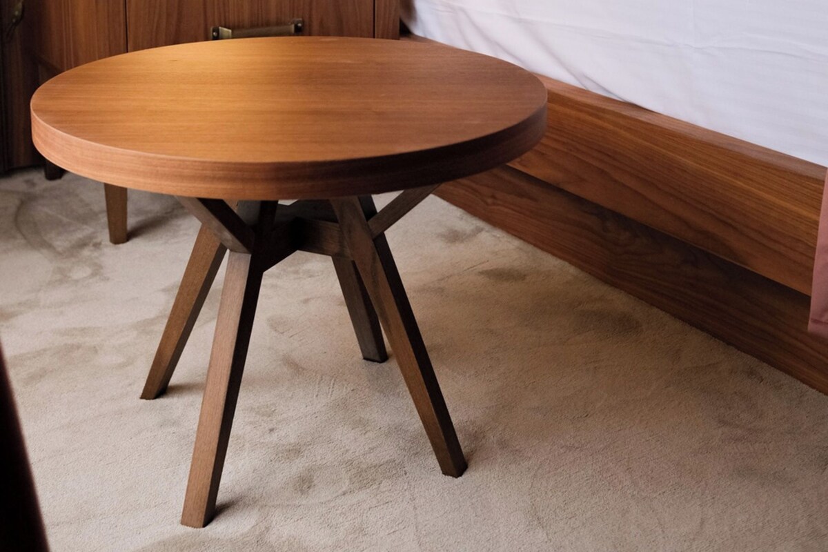 Une chambre d’hôtel avec une table de chevet en bois, une table ronde en bois, un lit avec une literie blanche et marron, et un papier peint à motifs.