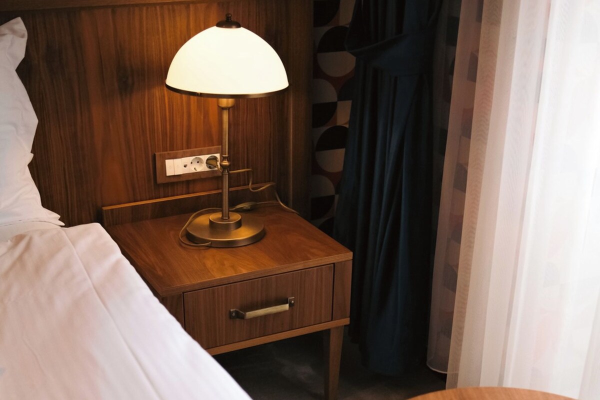 Un lit bien fait, avec une couette blanche et bleue, une table de chevet avec une lampe et une petite table ronde en bois.