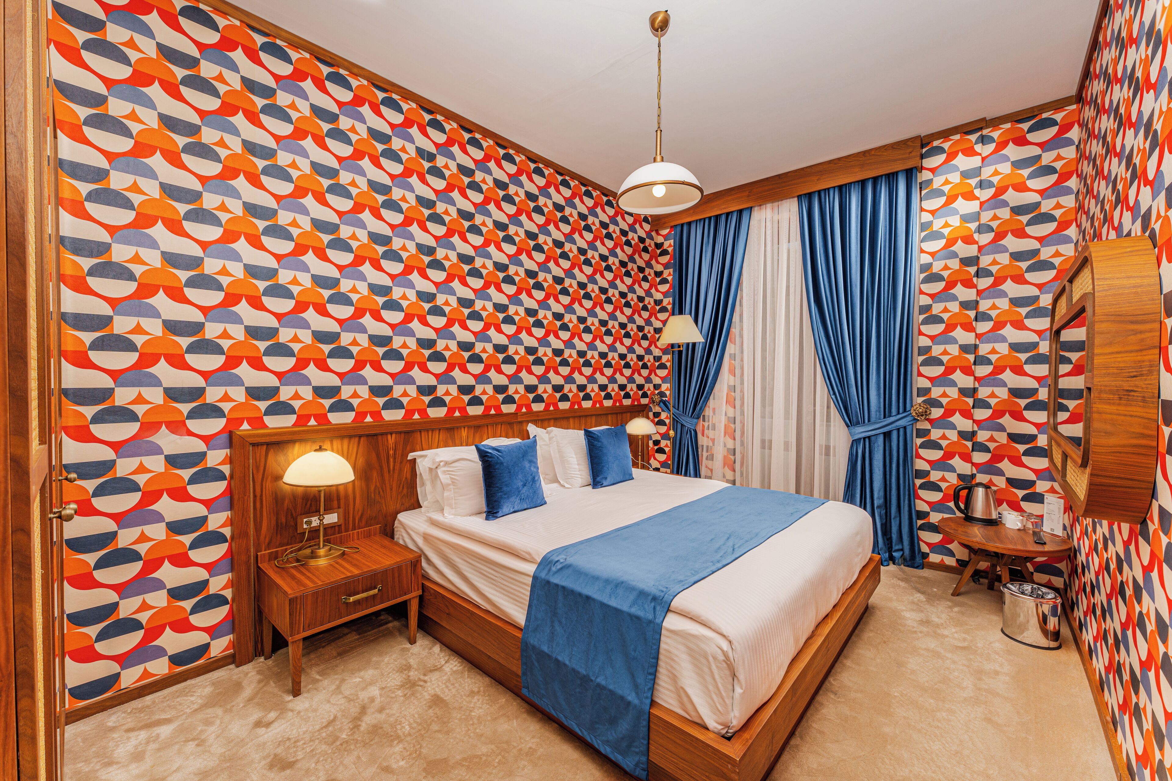 Una habitación de hotel con papel tapiz estampado, una cama con manta azul, una lámpara de noche, una mesita de madera y una pequeña mesa con un hervidor.