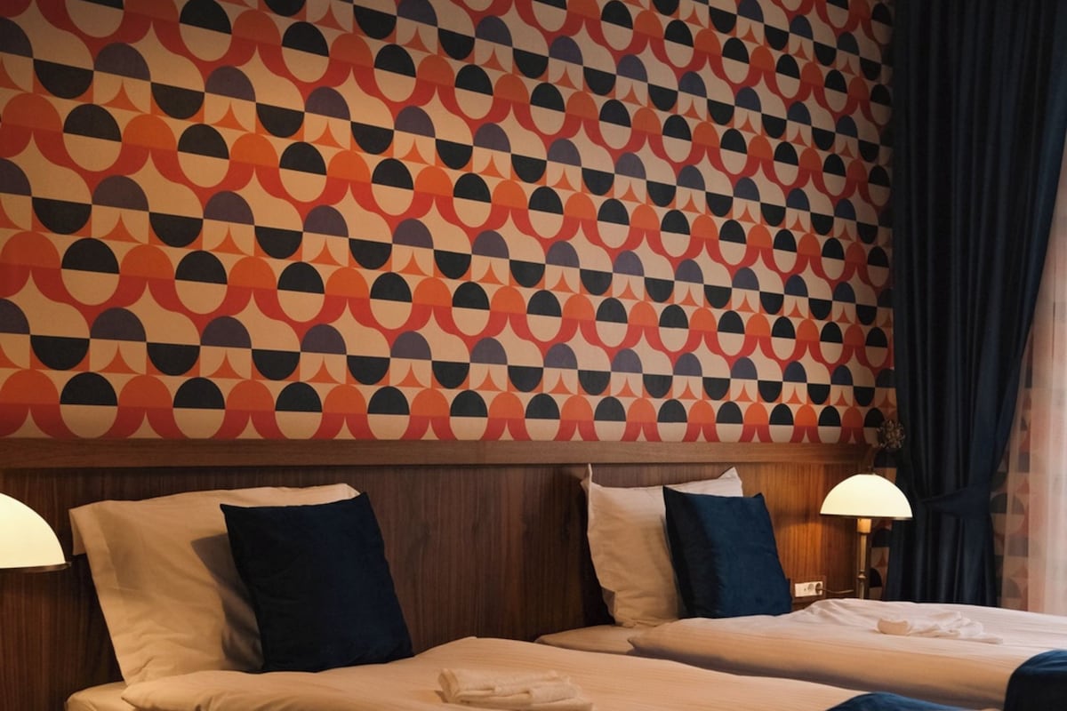 A hotel room with two beds, a patterned wall, and a nightstand with a lamp.