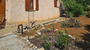 A rustic building with a stone facade, wooden shutters, and a gravel pathway leading to a garden area with rocks and plants.