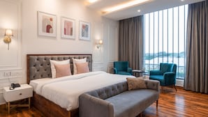 Una habitación de hotel moderna con una cama grande, un sofá gris, dos sillones y vistas de la ciudad a través de amplias ventanas.
