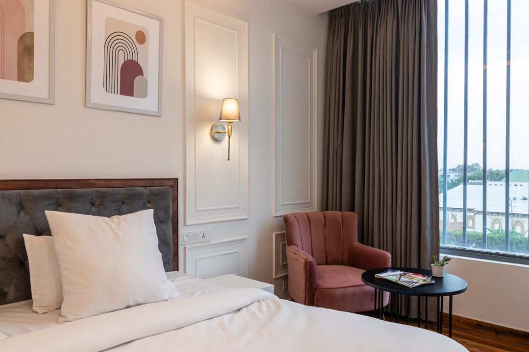 A hotel room with a bed, a chair, a small table with a magazine, and a window with curtains.